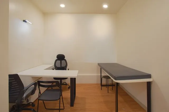 A clinic room with a desk and examination bed against pale walls.